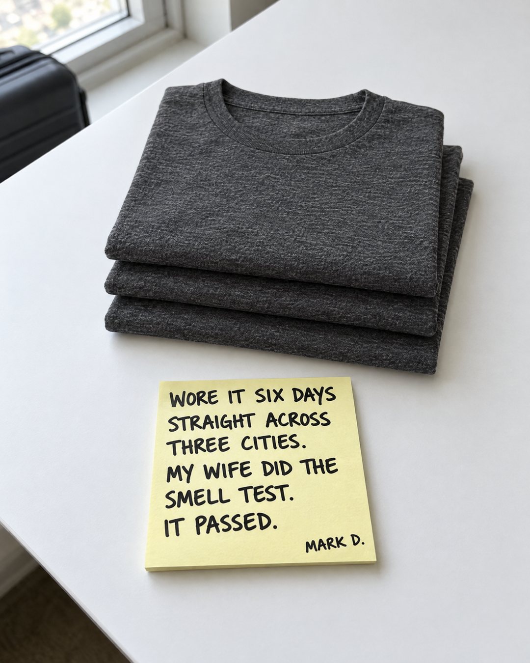 Three folded charcoal merino T-shirts stacked on a hotel desk, with a handwritten sticky note: "Wore it six days straight across three cities. My wife did the smell test. It passed." Mark D.